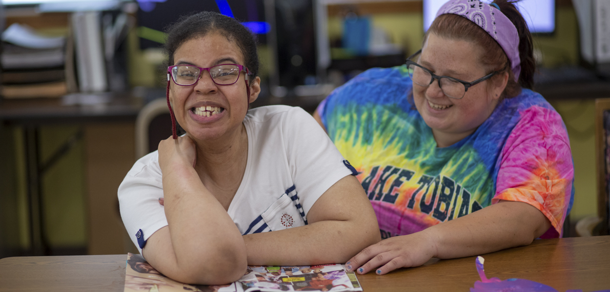 Two women sitting at desk. Client is smiling, team member is sitting next to her helping her read a magazine.