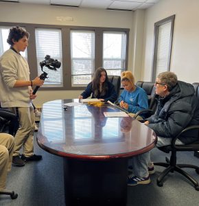 Three people talking at a table with a boy recording them with a camera.