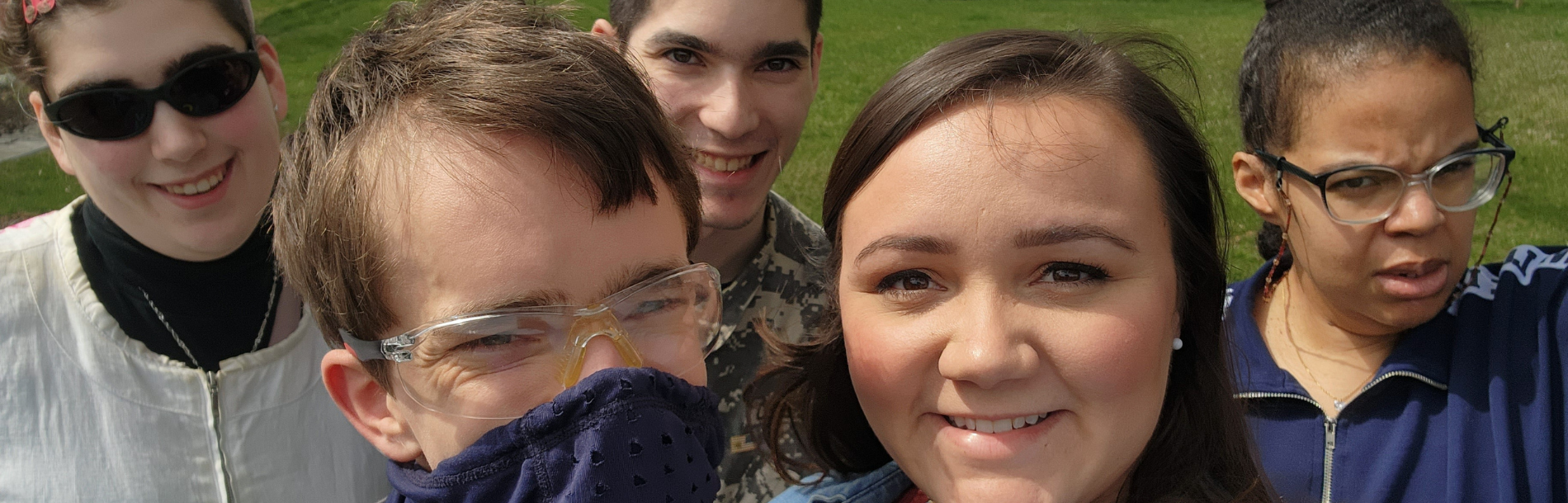 5 people smiling at camera while they take a group selfie outside.