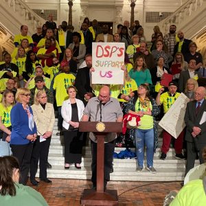 A man standing at a podium with a group of people advocating for DSP wages behind him.