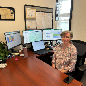 Kelly sits at her desk in her office with three monitors and a laptop open on her desk.