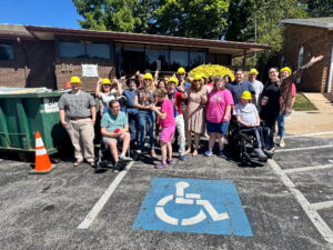 A group of people (some wearing hardhats) stands in front of a building in a parking lot with excited expressions on their faces.