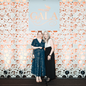 Jenn Hobbs and Kari Ebeling smile and pose together in front of a Gala backdrop.