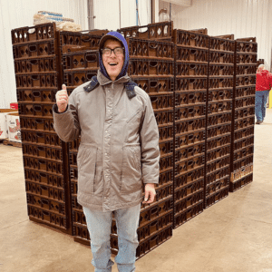 Mark smiles at the camera and holds a thumbs up as he stands in front of a large stack of pallets that's almost taller than he is.
