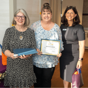 Jen Skelly (center) smiles and holds up her award certificate and poses with two other women.