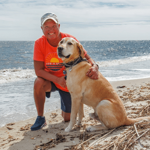Melanie Svitich and a yellow lab pose on the beach with the ocean in the background.