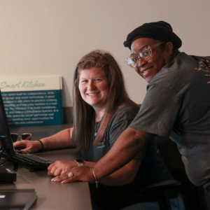 Two women smile and lean over a computer together.