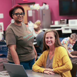 Two women pose next to each other, one sitting at a table and one standing beside her. 