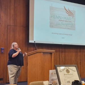 Allis speaks into a microphone at a podium with a screen in the back that shows the Penn-Mar logo.
