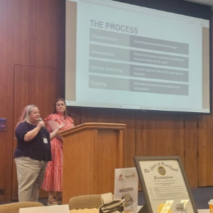 Allis speaks into a microphone at a podium with someone standing next to her. A screen in the back shows a slideshow titled "The Process."